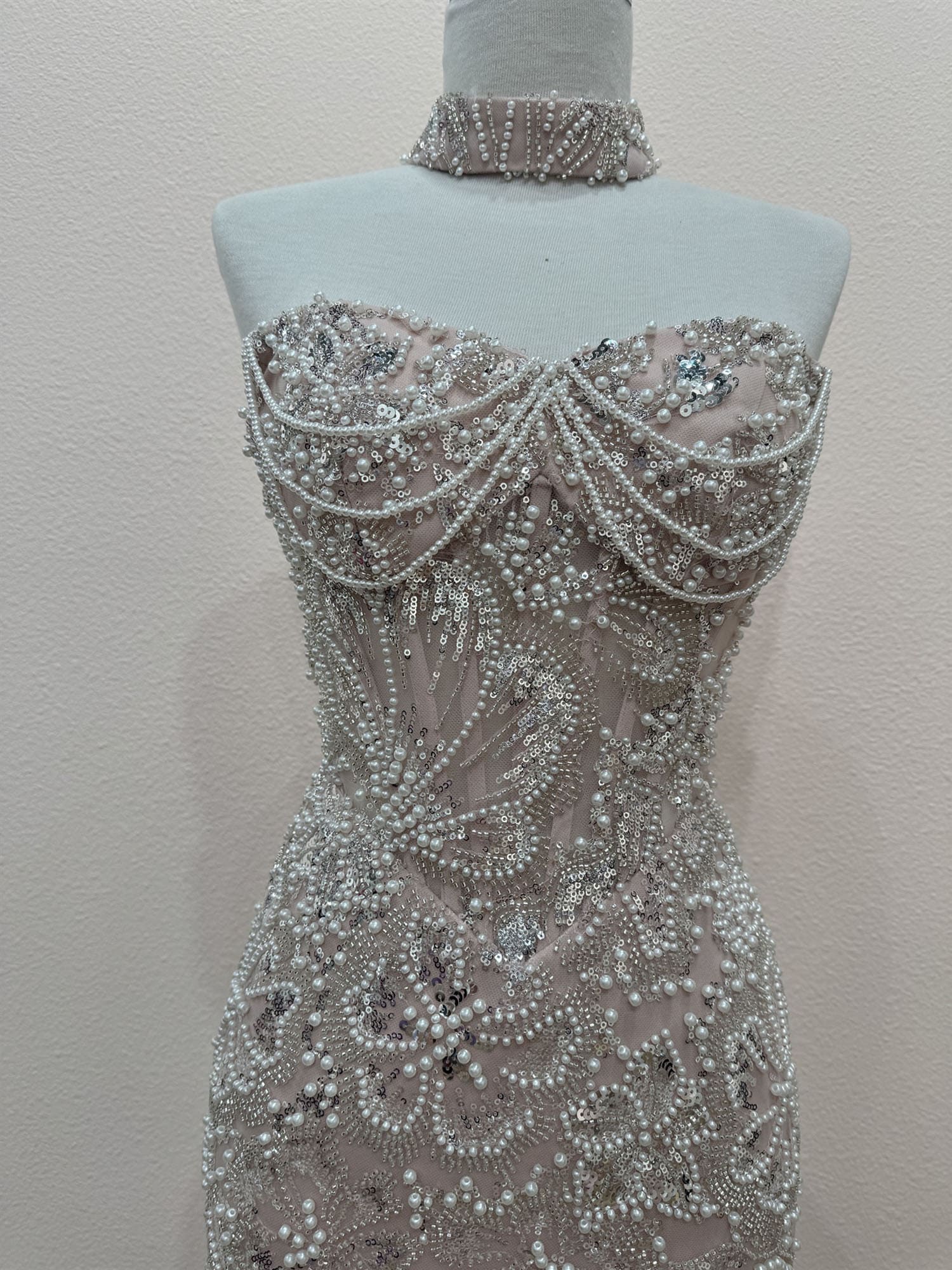 A strapless fitted gown displayed on a mannequin torso, covered in dense beadwork and pearl-like embellishments. The dress has a sheer blush-beige base with silver-toned beading and a matching beaded choker collar.