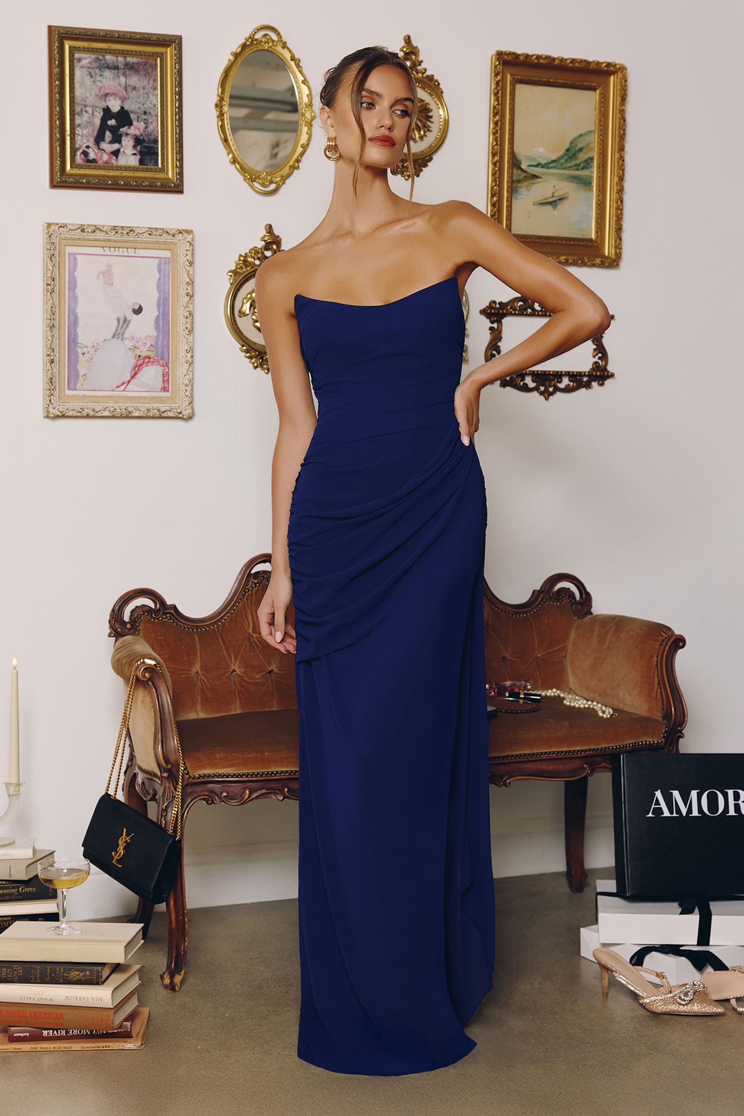 Woman in a blue strapless gown standing in an elegant room with a wooden bench and framed pictures.