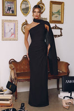 A woman models a floor-length black one-shoulder evening gown in a styled room with framed wall art, a bench, and shoes on the floor.