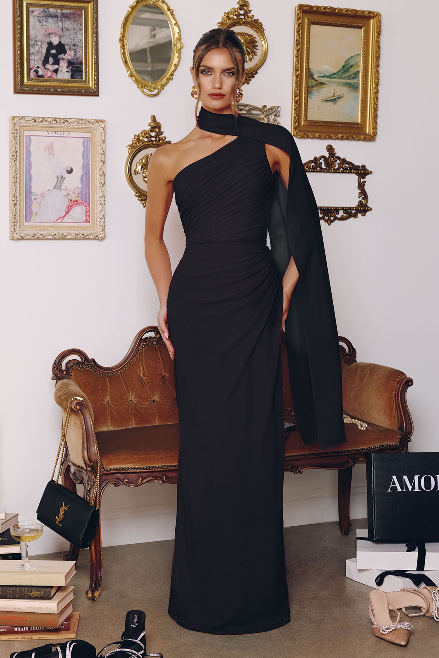 A woman models a floor-length black one-shoulder evening gown in a styled room with framed wall art, a bench, and shoes on the floor.