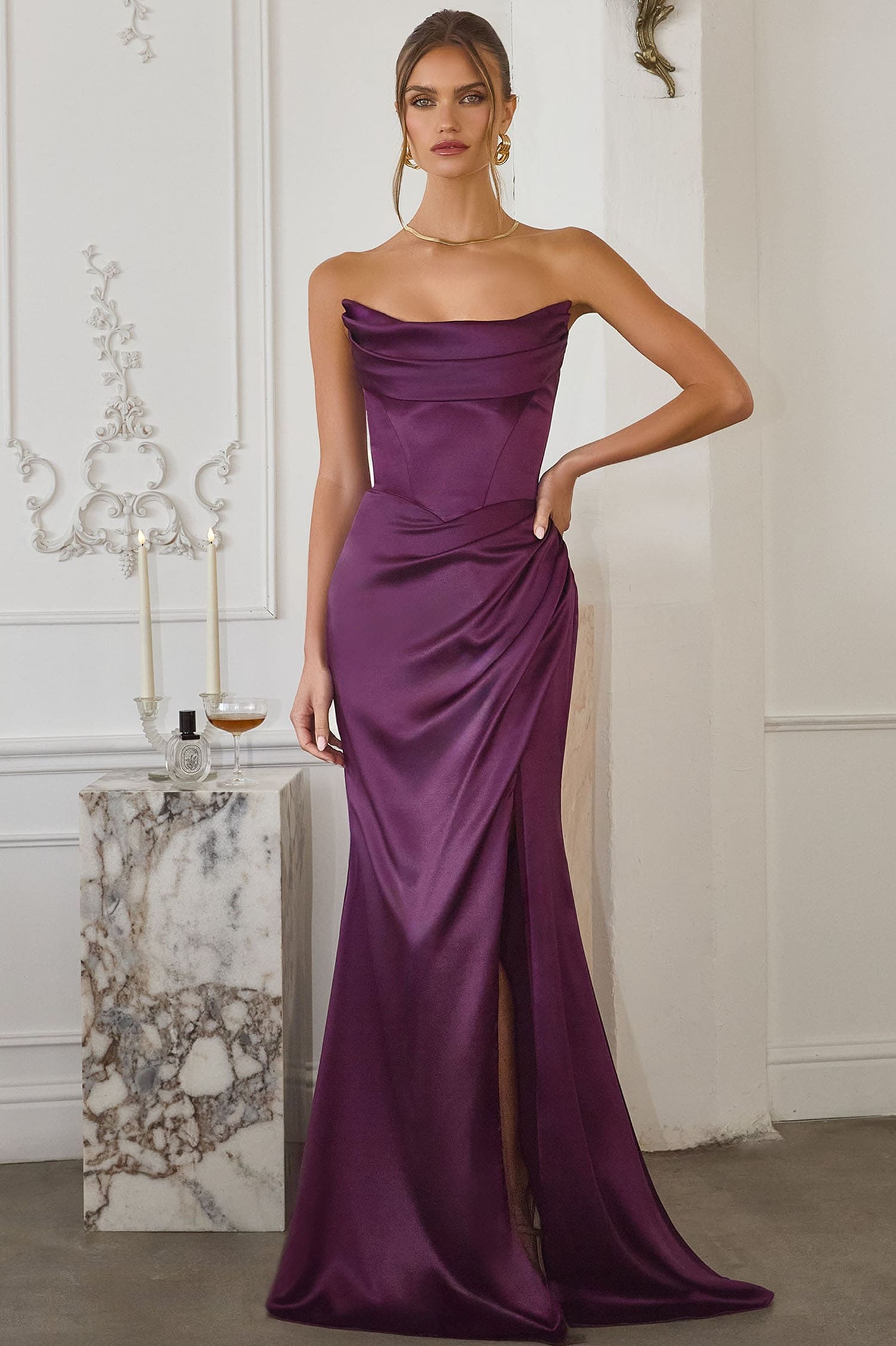 A woman models a strapless, floor-length purple satin gown with a draped bodice, gathered skirt, and thigh-high slit in an elegant indoor setting with a marble side table.