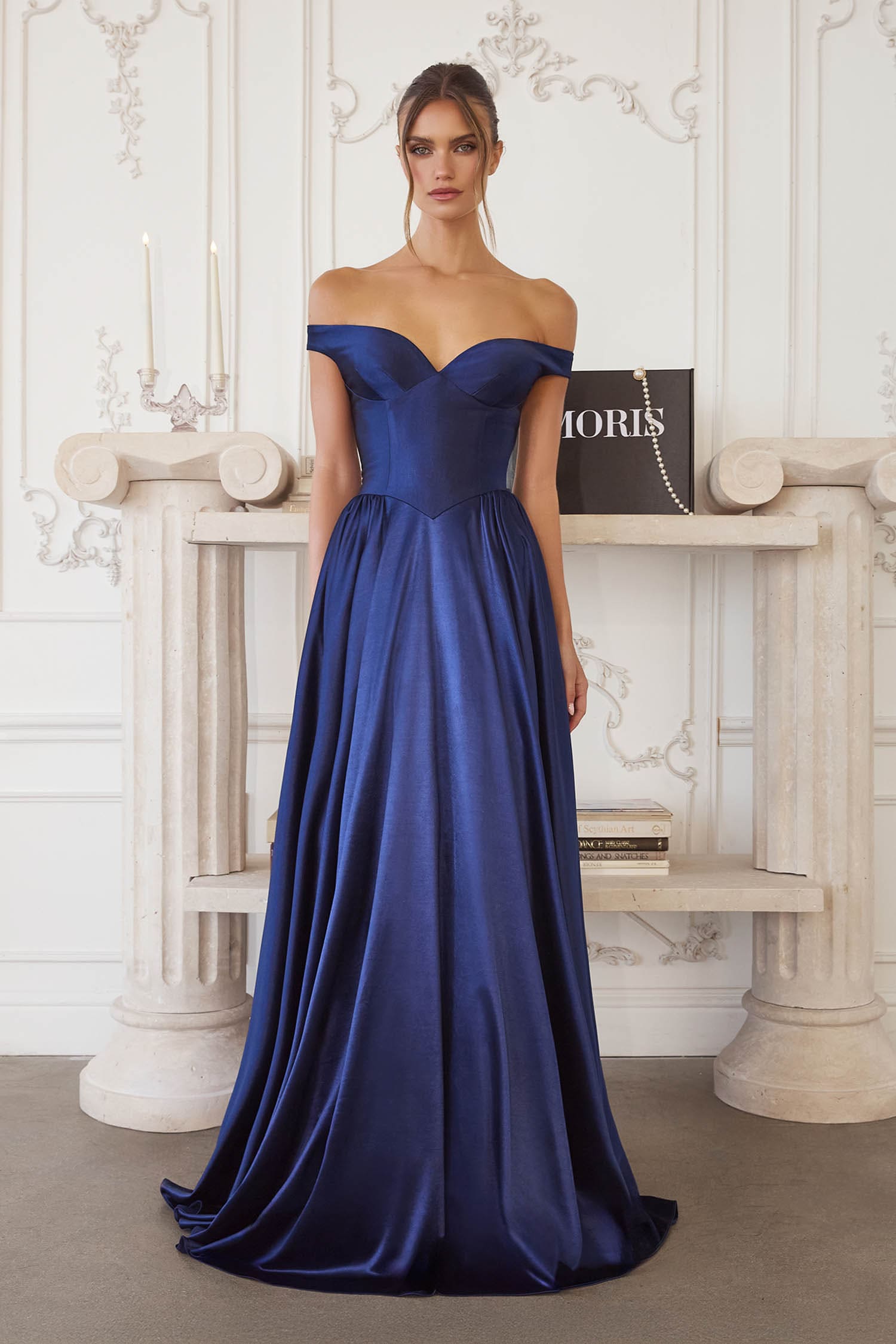 A woman stands in a grand interior wearing an off-the-shoulder floor-length satin gown in deep royal blue. The dress has a fitted bodice and full skirt, with a visible Dior bag and decorative console table behind her.