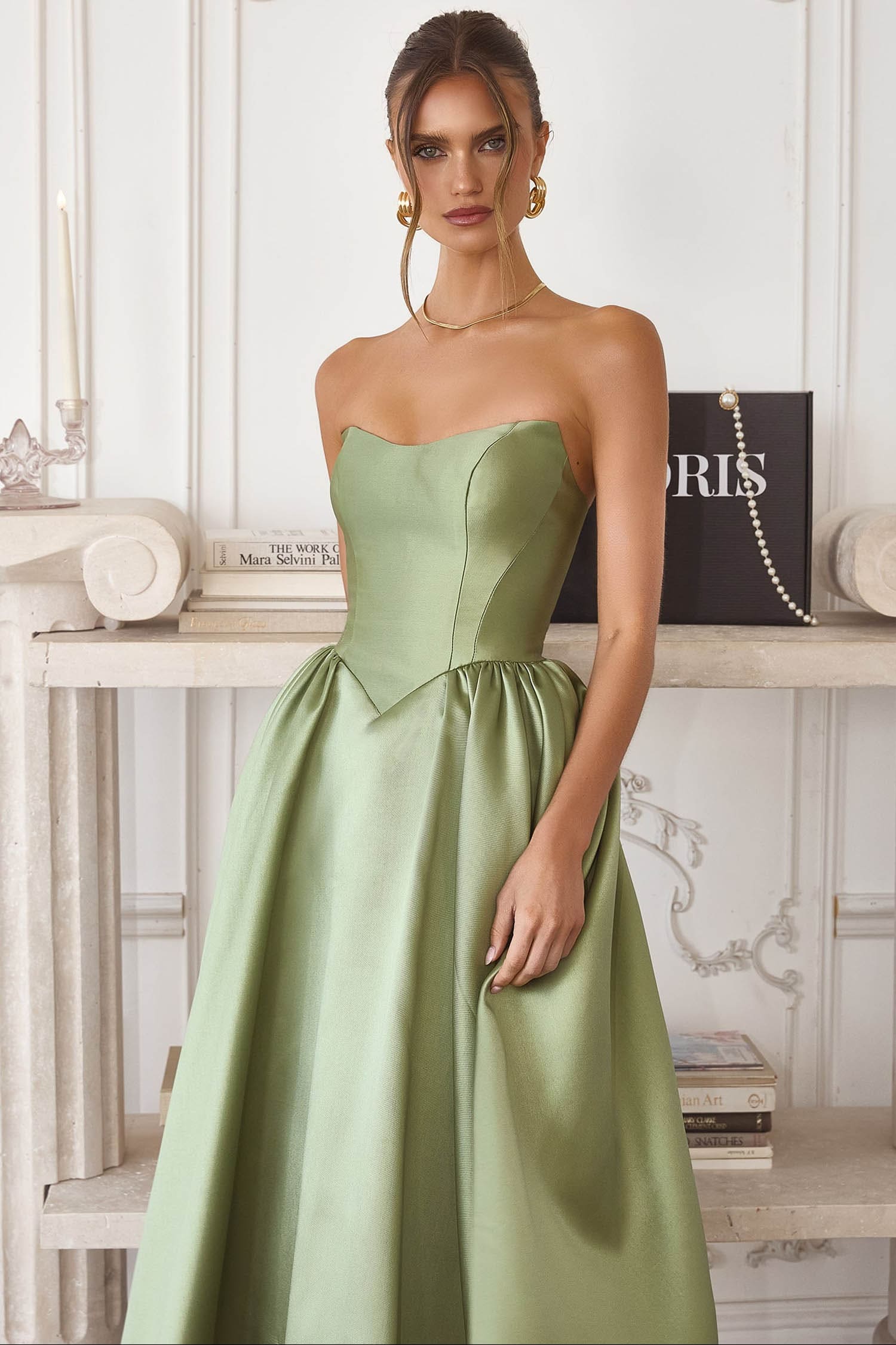 A woman models a strapless light sage green satin ball gown with a sweetheart neckline, structured corset bodice, and full A-line skirt, posed in front of a white paneled wall and console table.
