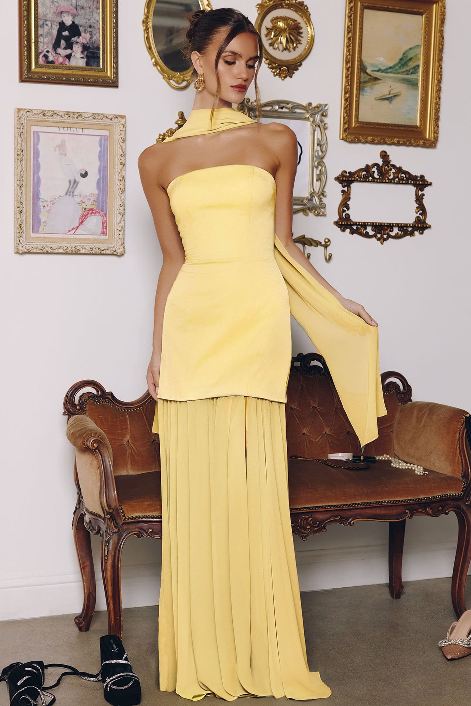A woman models a yellow one-shoulder strapless maxi dress in a vintage-style room with a brown leather bench and framed wall art.
