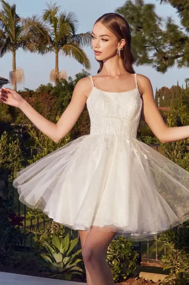 A woman in a short, white, fluffy dress twirls gracefully on a patio. She has long hair and is surrounded by lush greenery and palm trees under a clear sky.