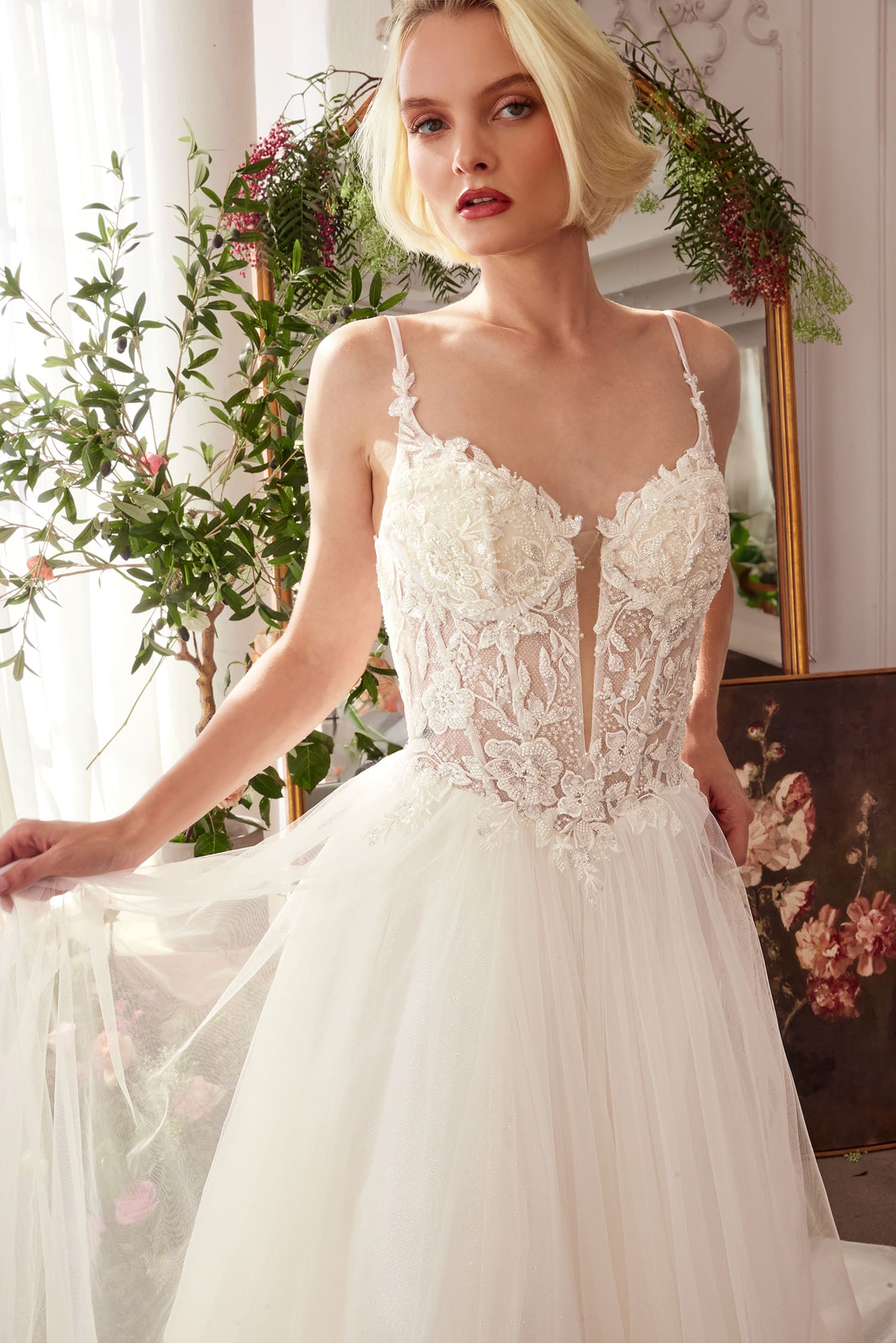 Woman wearing a white lace wedding dress in a decorated room with plants and artwork.