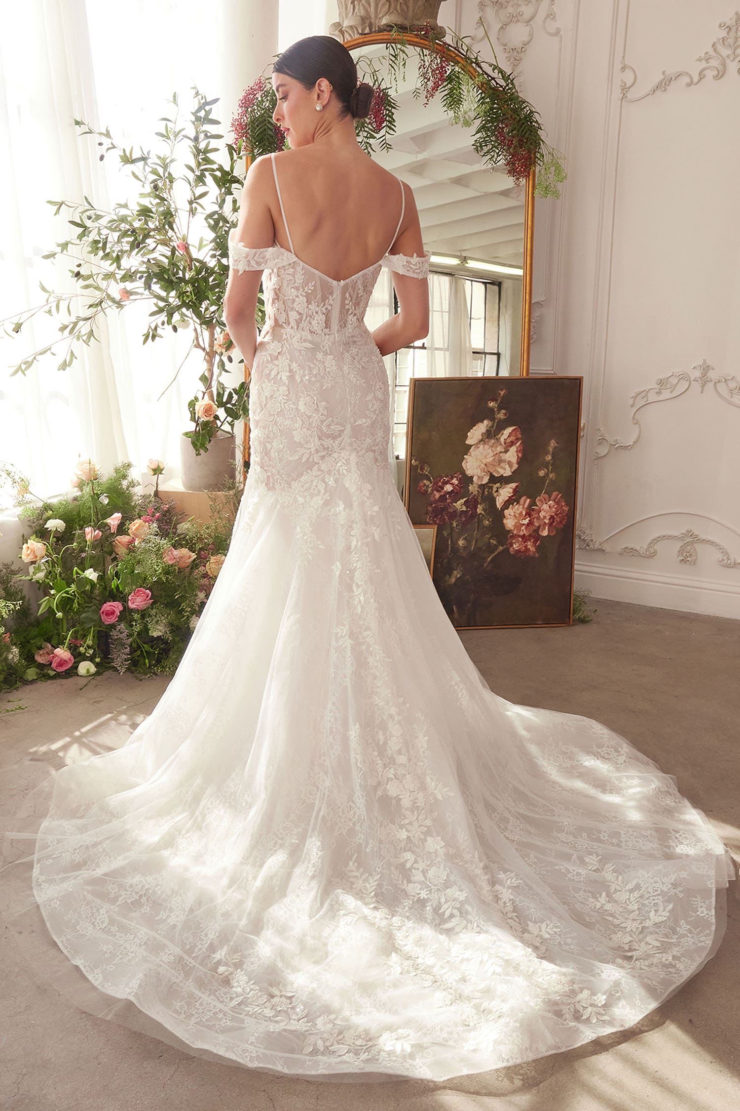 Woman in a white lace wedding dress standing in a decorated room with flowers and a mirror.