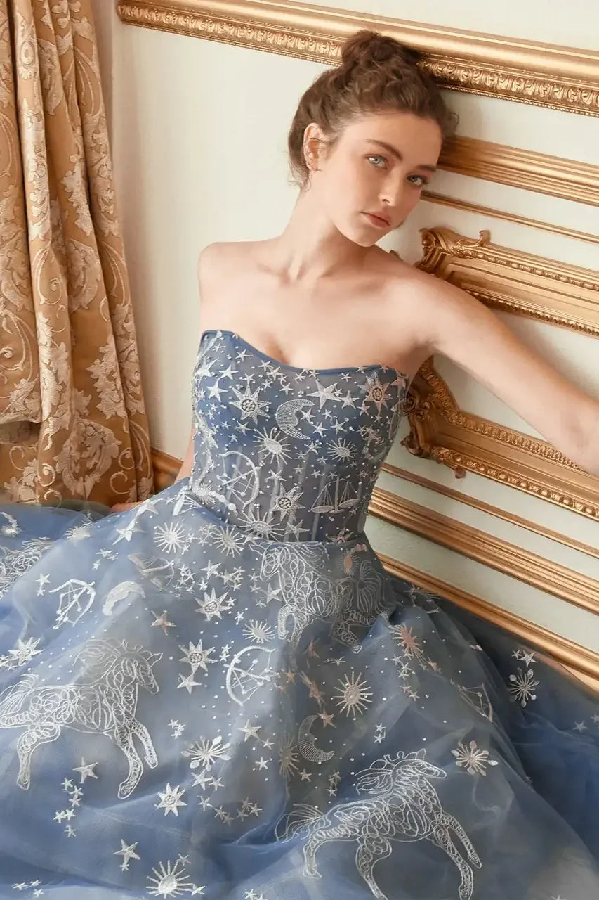 A young woman poses against a wall, wearing a strapless, intricately embroidered blue gown adorned with celestial designs, including stars and unicorns, while drapes hang in the background.