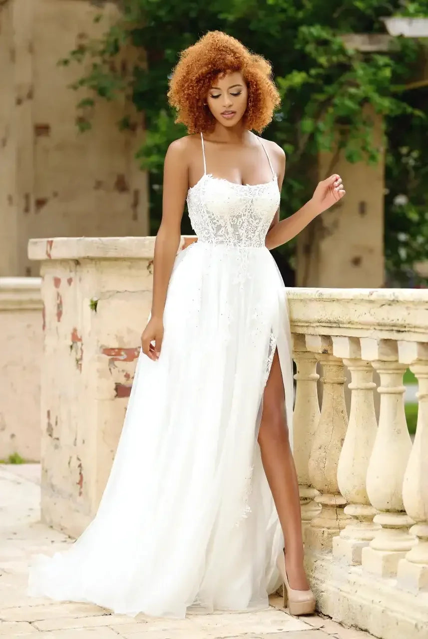 A woman in a flowing white gown with lace details stands gracefully beside a decorative stone railing. Natural greenery surrounds her, creating a serene, elegant atmosphere.