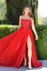 A woman in a flowing red gown walks gracefully, showcasing a leg slit. She is surrounded by lush greenery, with tall hedges framing the background.