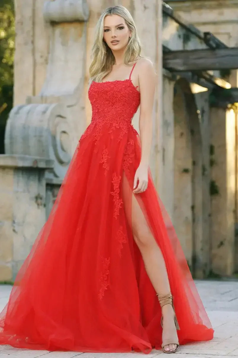 A woman stands confidently in a sweeping red gown with a thigh-high slit and lace detailing. She poses against a backdrop of stone architecture and greenery.