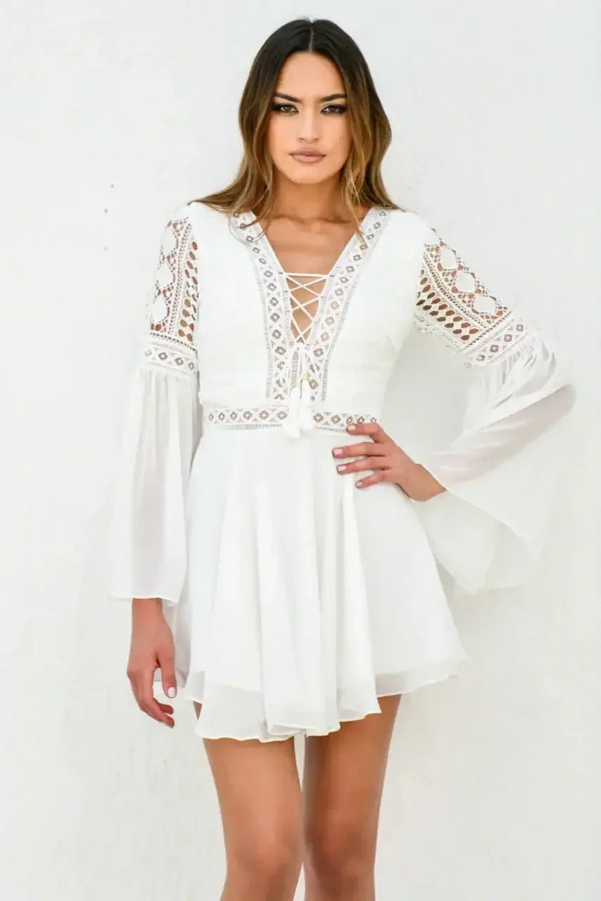 A woman poses confidently in a white, intricately designed dress with lace details and bell sleeves against a plain, textured white wall, showcasing a stylish, elegant look.