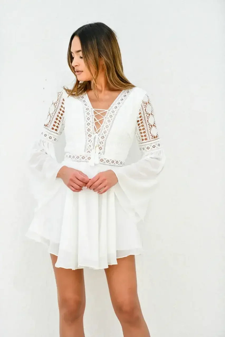A woman stands gracefully in a white, intricately designed dress with long, flared sleeves. She gazes down thoughtfully against a textured white wall, embodying elegance and serenity.