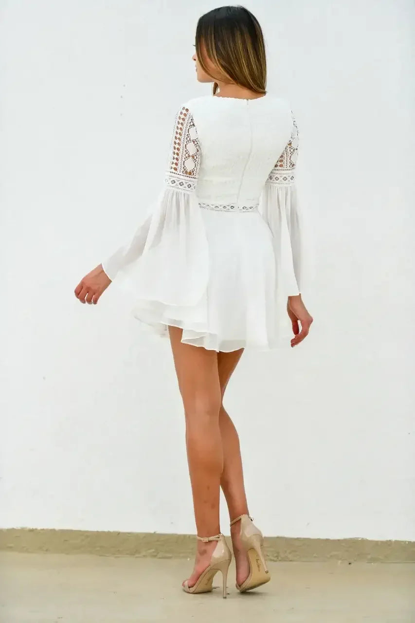 A woman in a white, flowing dress with long bell sleeves is walking away, revealing a textured bodice and her strappy, high-heeled shoes against a plain white wall.