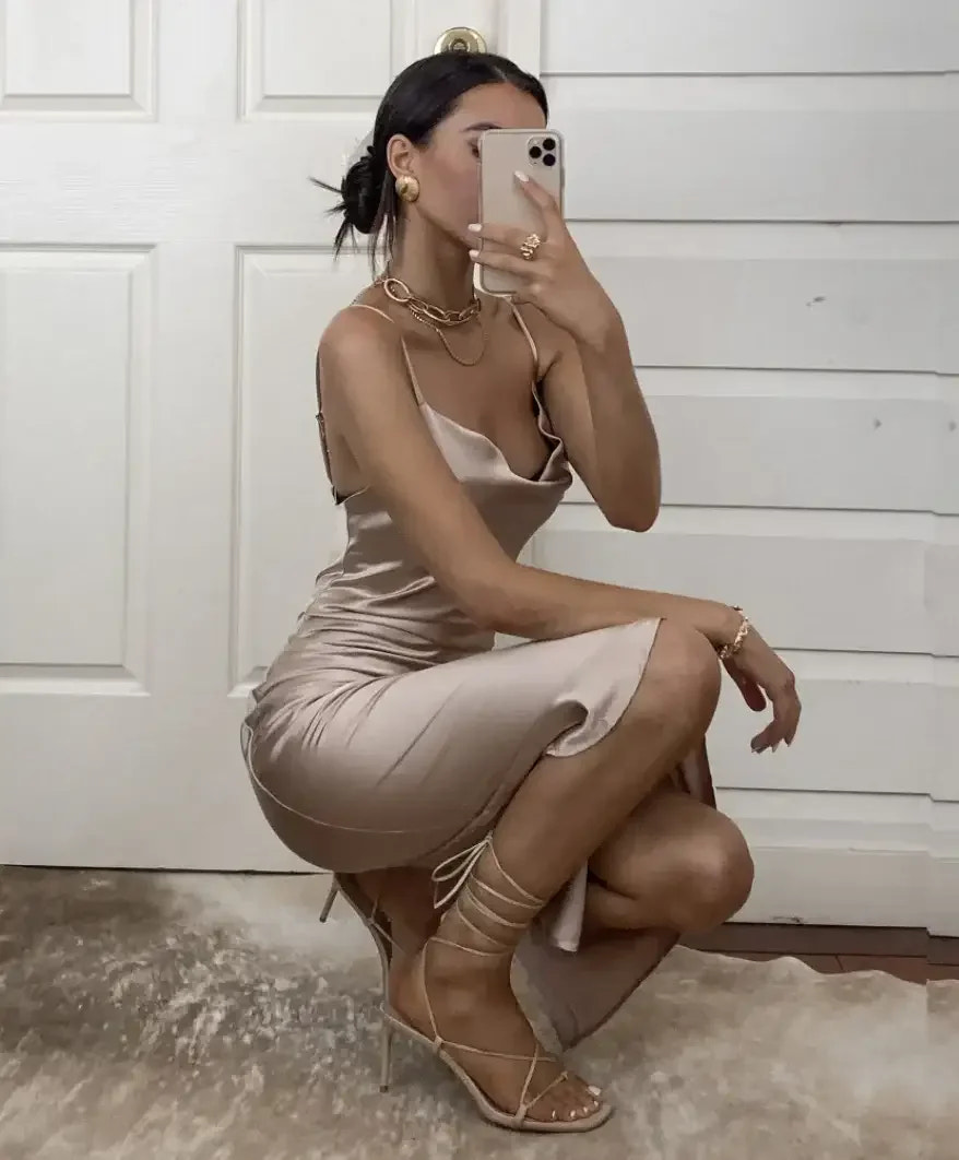 A person in a satin beige dress poses while taking a selfie. They are accessorized with jewelry, sitting on a rug in front of a light-colored door.