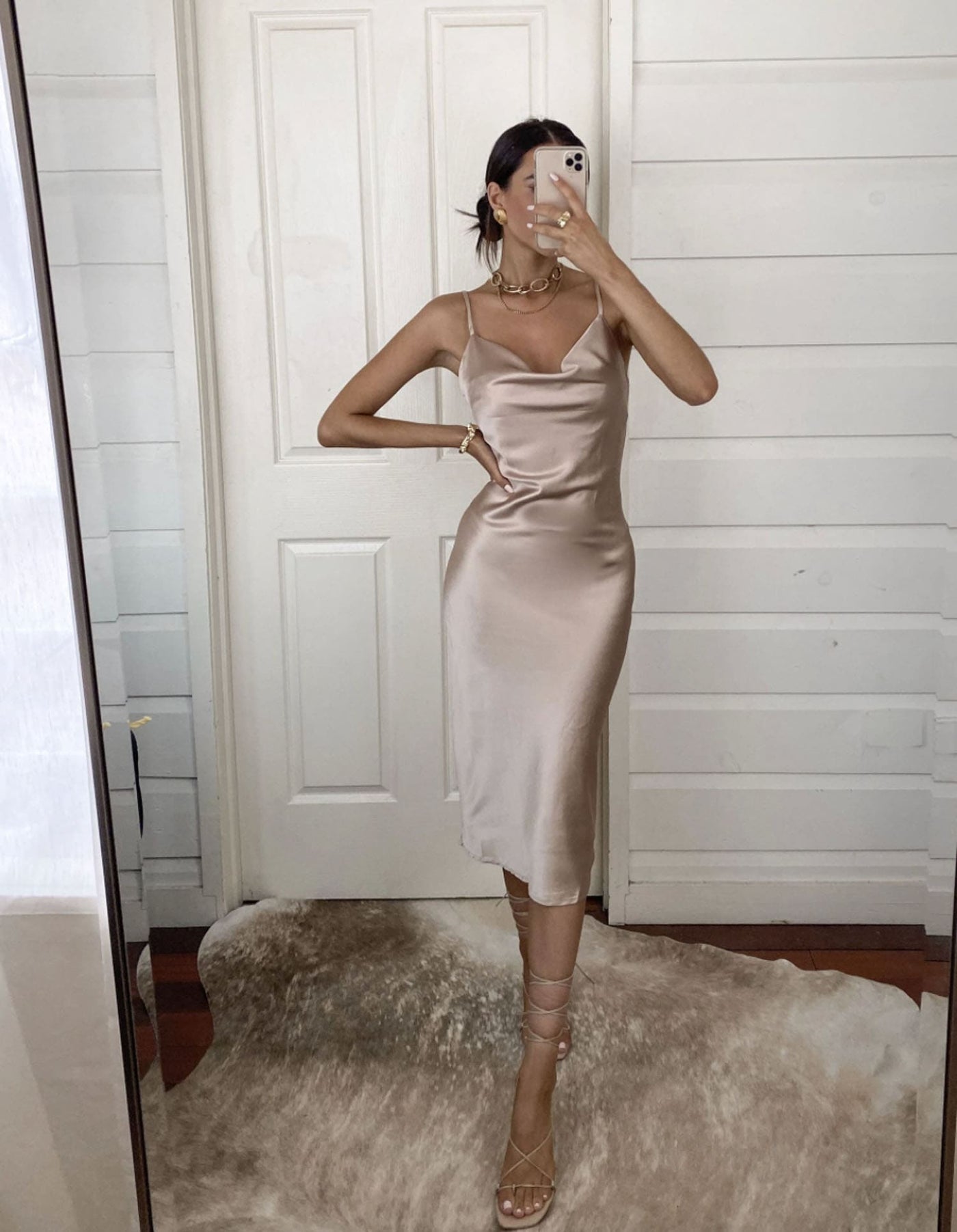 A person is posing in front of a mirror, wearing a light silk dress and strappy sandals. The background features wooden panels and a plush rug on the floor.