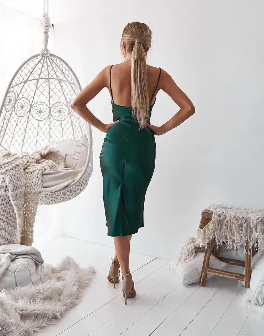 A woman wearing a green satin dress stands gracefully with her back to the viewer. She poses in a bright, neutral room featuring a hanging chair and soft dÃ©cor.