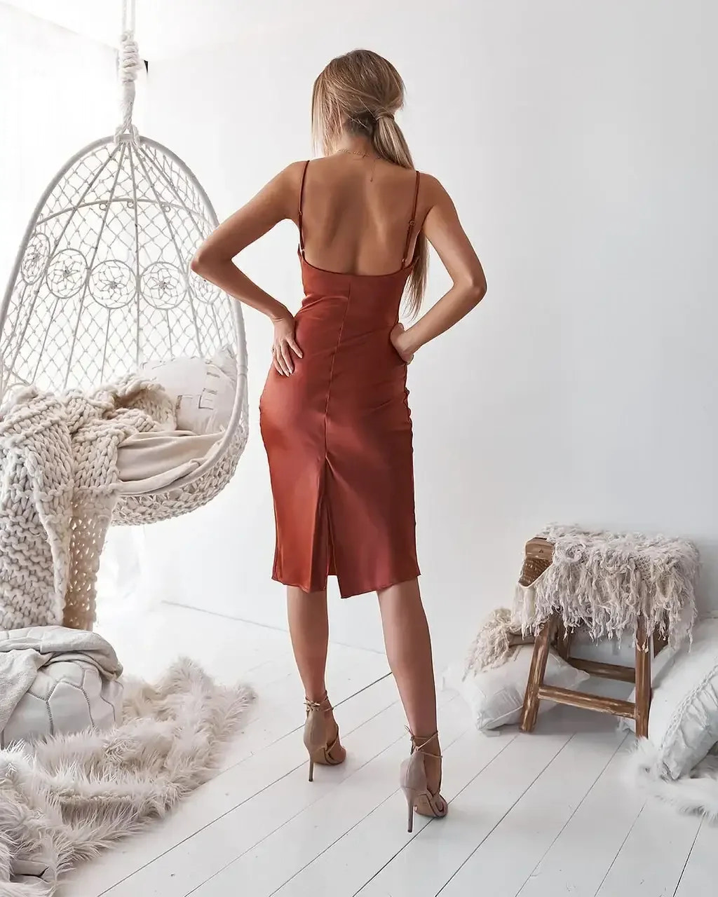 A woman in a smooth, rust-colored dress stands with her back to the viewer, hands on hips, in a bright room featuring a hanging chair and cozy textiles.