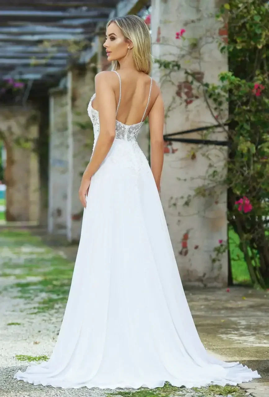 A woman in a flowing white gown stands with her back to the camera, showcasing intricate details. She is set against a backdrop of stone columns and vibrant greenery.