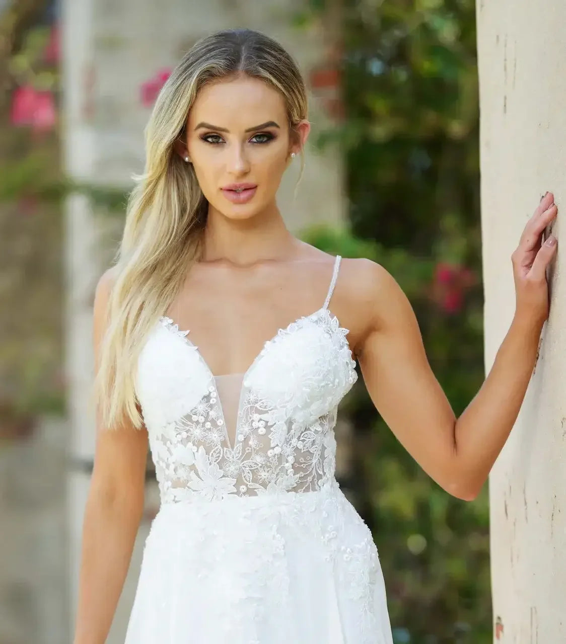 A woman in a delicate, lace-trimmed white gown stands confidently against a light-colored wall, with soft greenery and flowers visible in the blurred background, enhancing a romantic atmosphere.