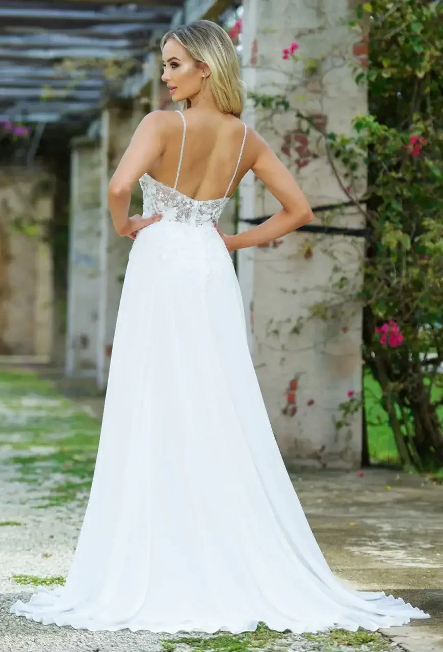A woman stands in a flowing white dress with delicate lace detailing, showcasing her back. She is positioned outdoors against a backdrop of stone pillars and greenery.