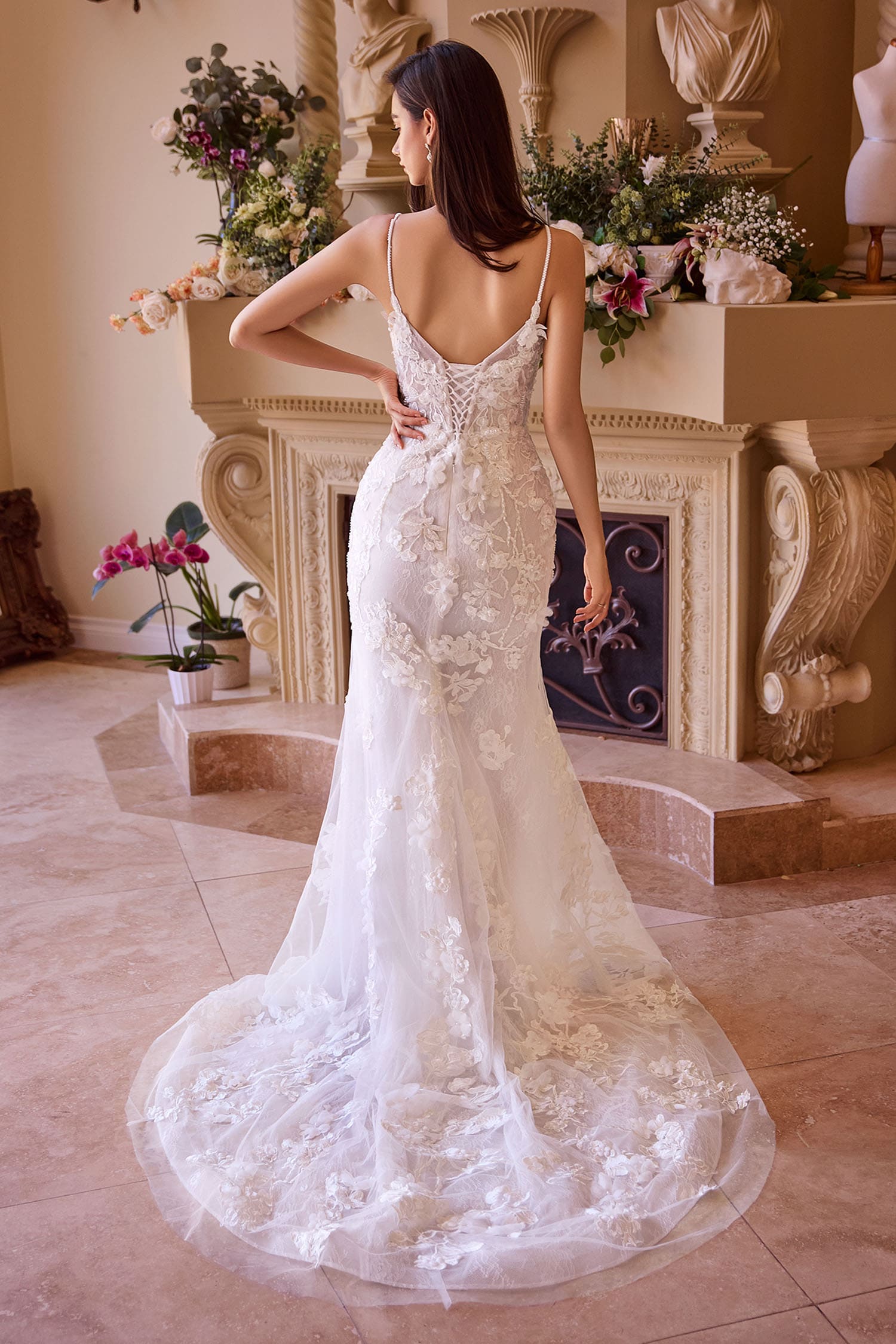 Woman in a white lace wedding dress standing in a decorated room with flowers and a fireplace.
