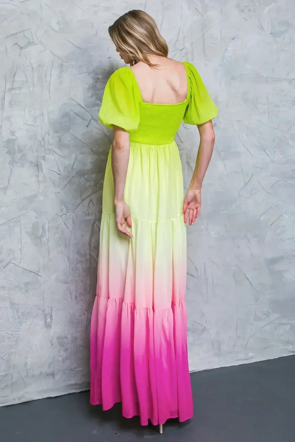 A woman stands with her back facing a textured grey wall, wearing a vibrant gradient dress that transitions from bright yellow to pink, featuring puffed sleeves and a fitted bodice.