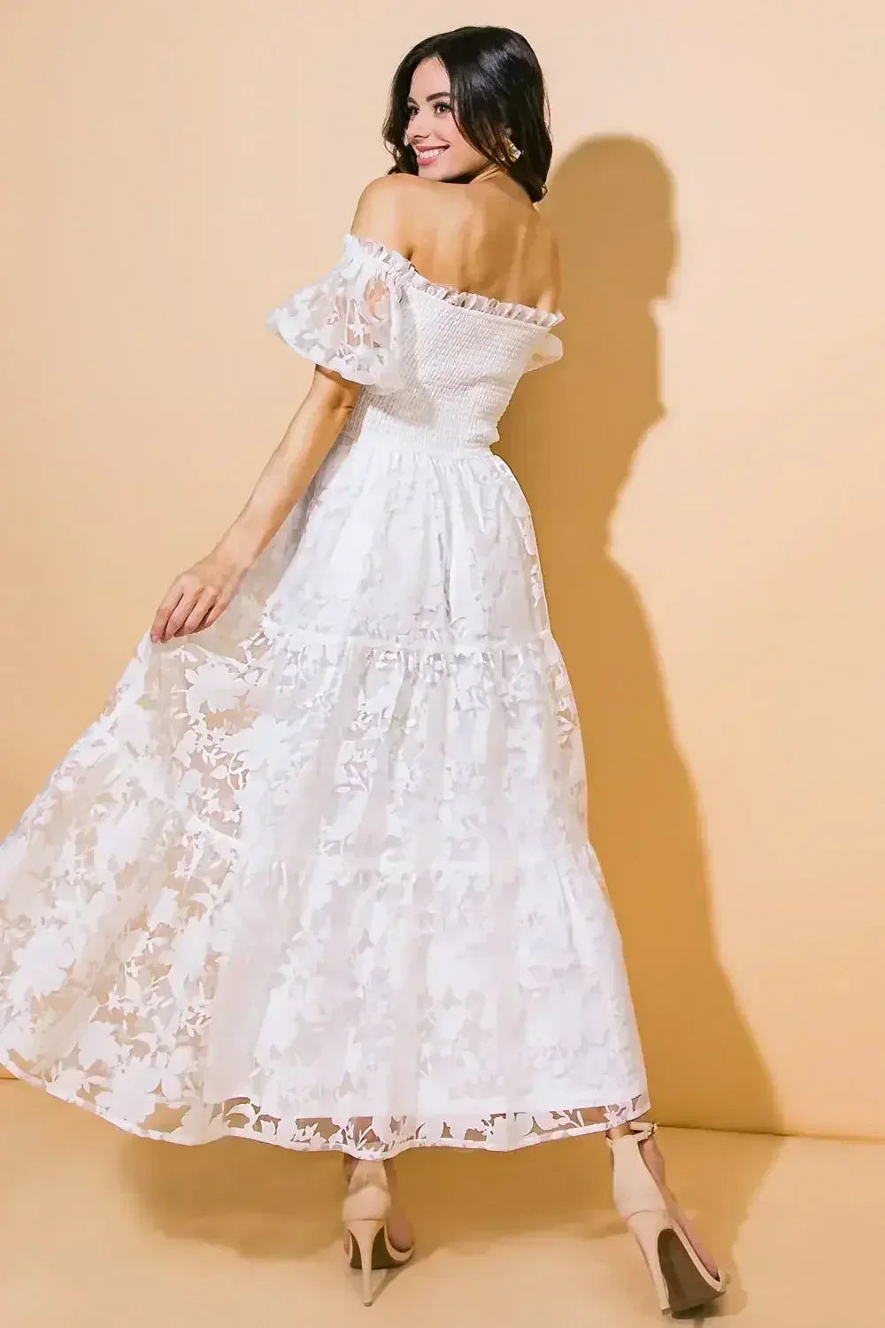 A woman in a flowing white off-shoulder dress playfully turns, showcasing intricate floral patterns. The soft beige background enhances her cheerful expression and elegant style.