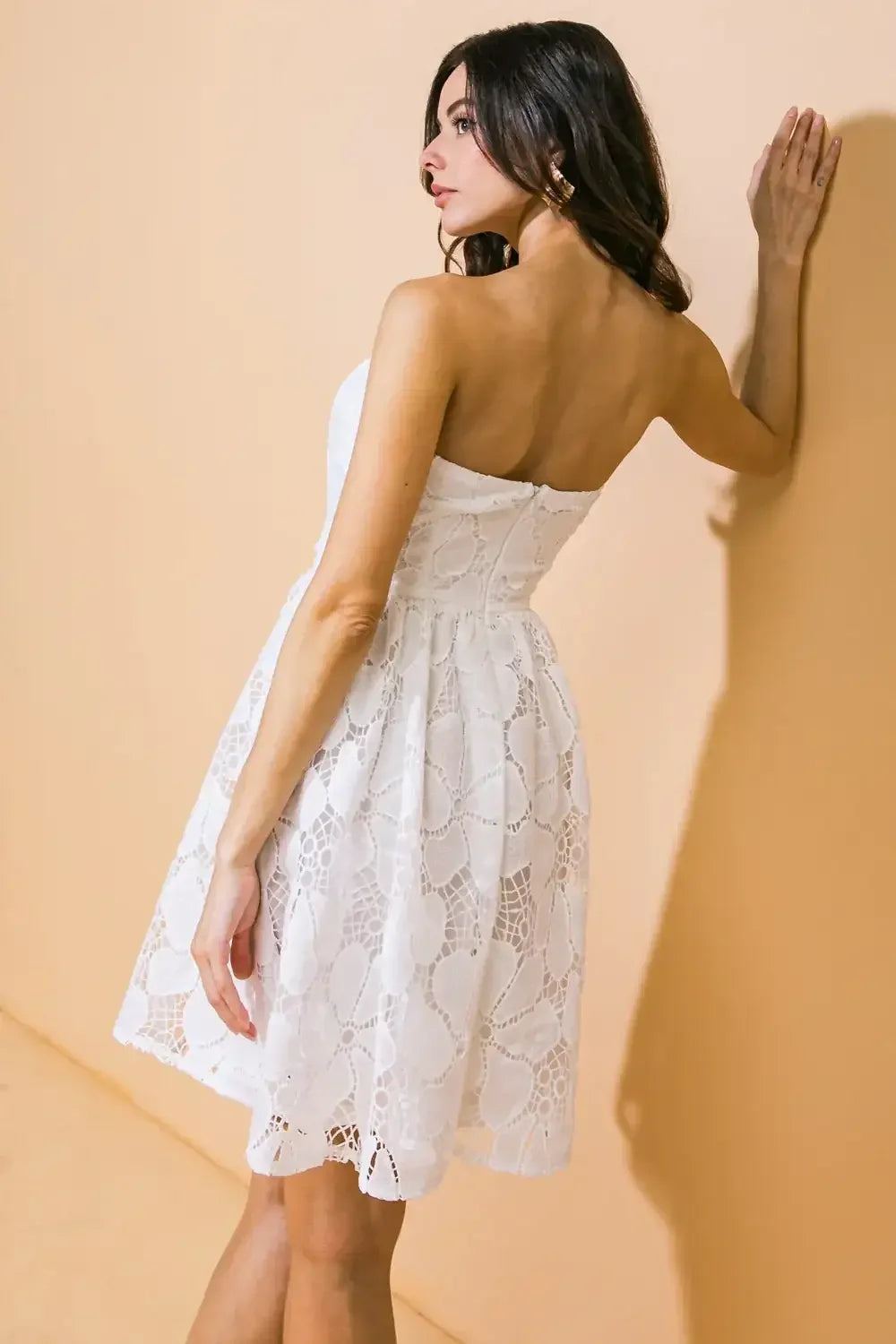 A woman is standing sideways, wearing a strapless white lace dress, with one hand resting on a light peach wall, her hair cascading down her back.