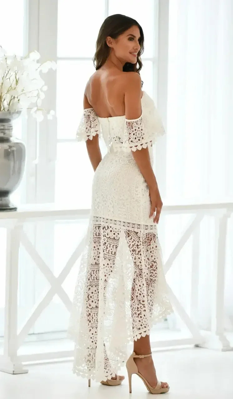A woman stands gracefully in a white lace dress with off-the-shoulder sleeves, displaying intricate patterns. She is positioned near a bright window, surrounded by minimal decor.