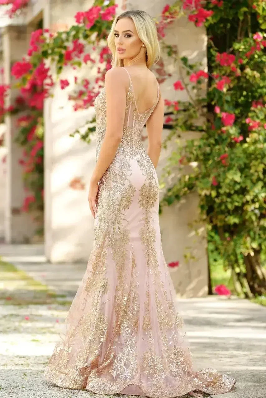 A woman in an elegantly fitted, gold-embroidered gown poses with her back to the camera, surrounded by blooming pink flowers and stone architecture in a sunny outdoor setting.