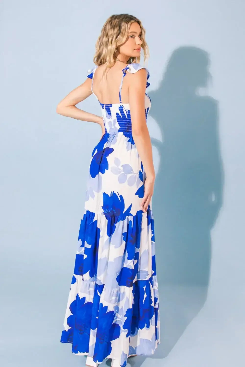 A woman in a floral blue-and-white maxi dress stands with her back to the camera, looking over her shoulder against a light blue background, showcasing the dress's ruffled details.