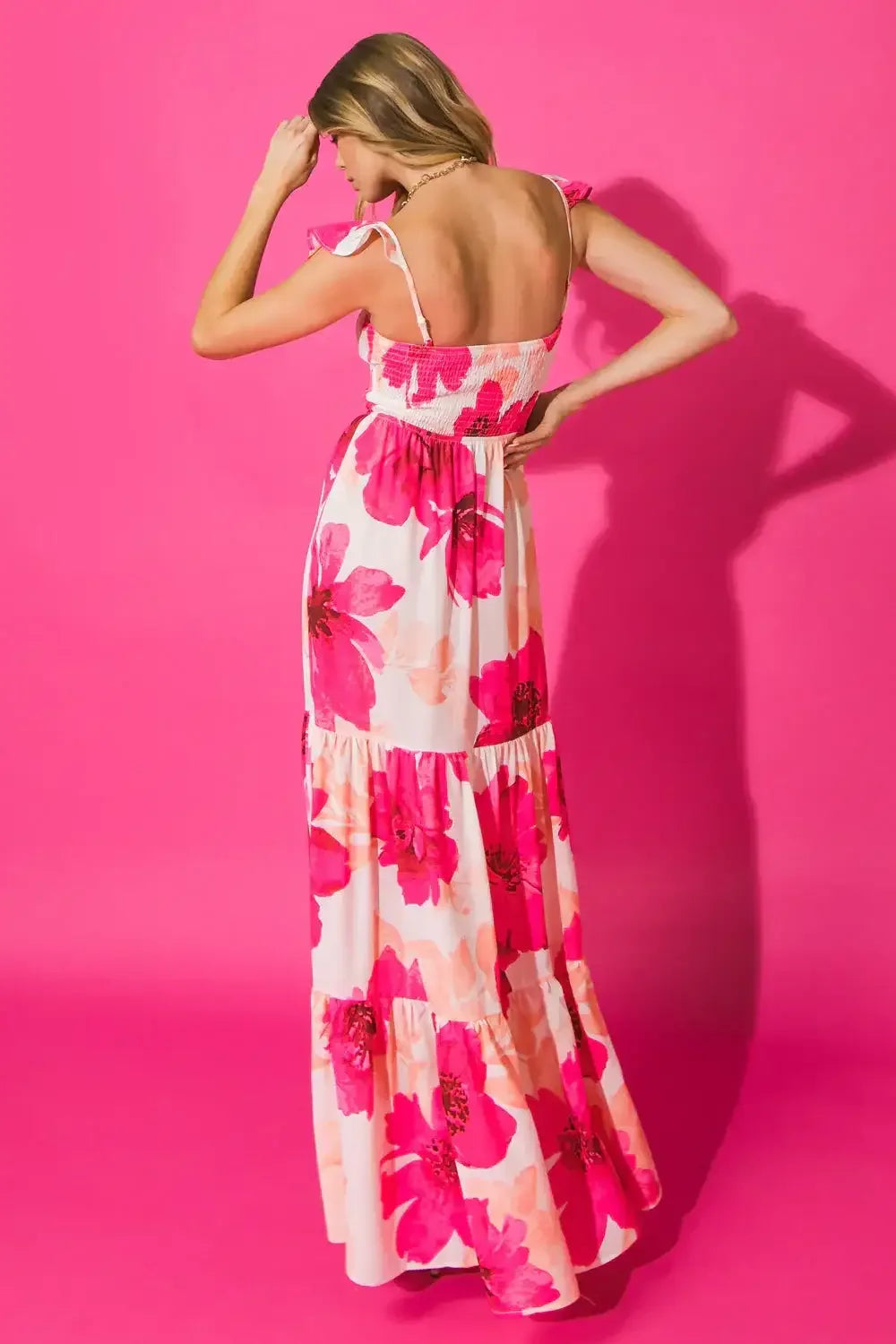 A woman stands wearing a long, floral-patterned dress with pink flowers. She touches her hair, facing away from the camera, against a bright pink background, creating a vibrant, stylish look.