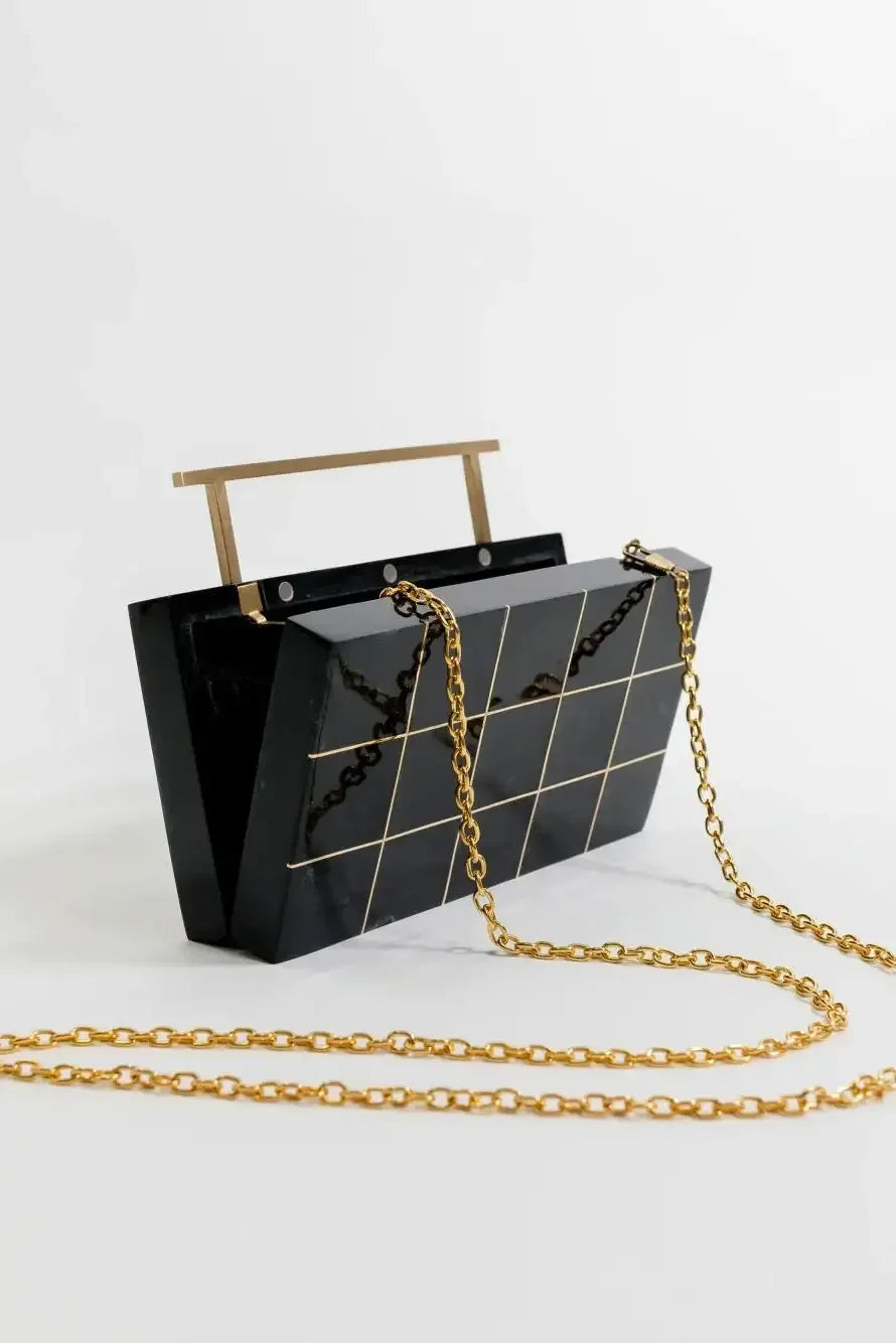 A black, rectangular handbag with gold chain strap features grid-like patterns. It stands upright on a plain white surface, with a horizontal handle at the top.