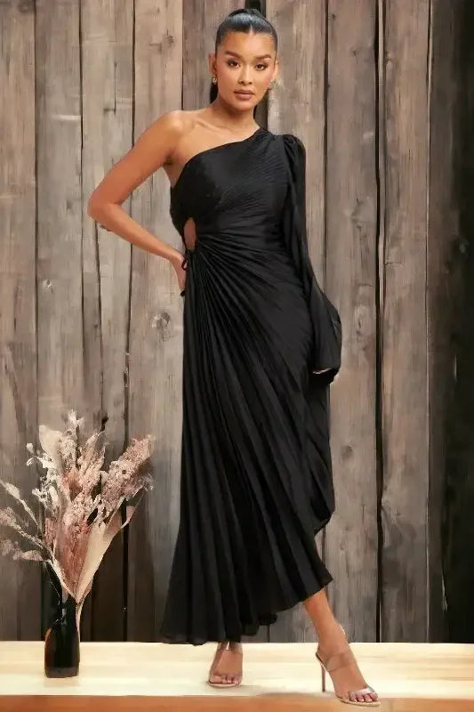 A woman stands confidently in a flowing, black, one-shoulder dress with pleats, showcasing a unique cutout. She poses against a backdrop of textured wooden panels, surrounded by elegant dried floral arrangements.