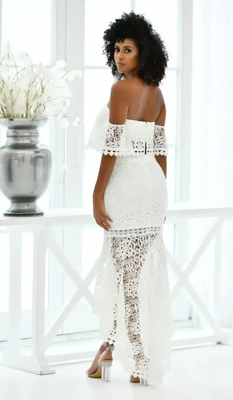 A woman with curly hair poses, wearing a white off-shoulder lace dress that features intricate floral patterns. She stands by a large window, with a vase of white flowers nearby.