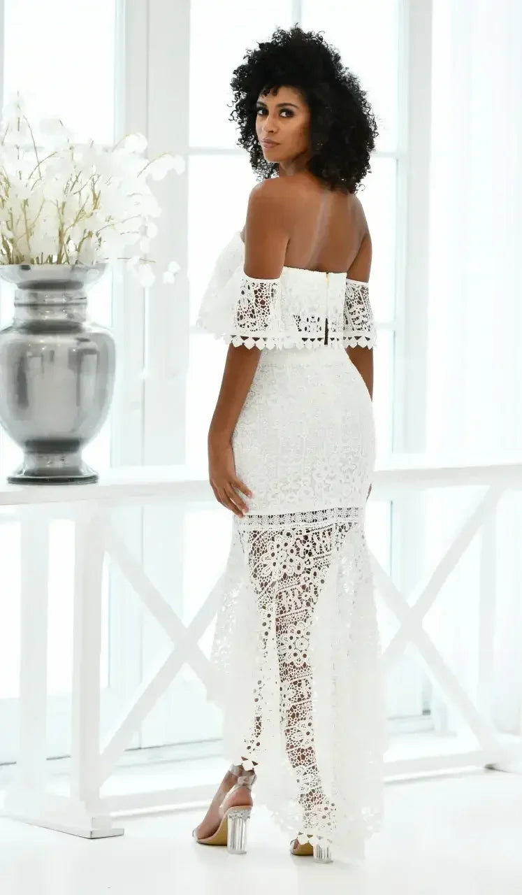 A woman with curly hair poses, wearing a white off-shoulder lace dress that features intricate floral patterns. She stands by a large window, with a vase of white flowers nearby.