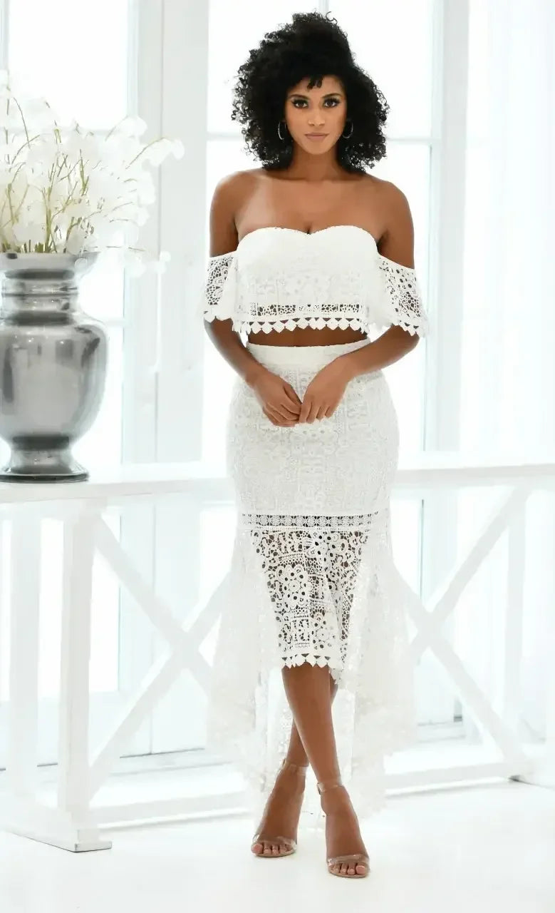 A woman stands confidently, wearing a white lace two-piece outfit with an off-shoulder top and a high-low skirt, in a bright, spacious room with large windows and a decorative vase nearby.