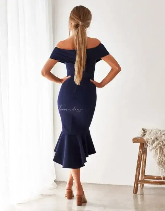 A woman wearing a fitted, navy off-shoulder dress stands with her back to the camera, posing in a well-lit room with white curtains and a wooden chair.
