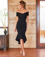 A woman stands gracefully in a fitted black dress with an off-shoulder design and a flared hem, inside a room with warm stone walls and large windows.