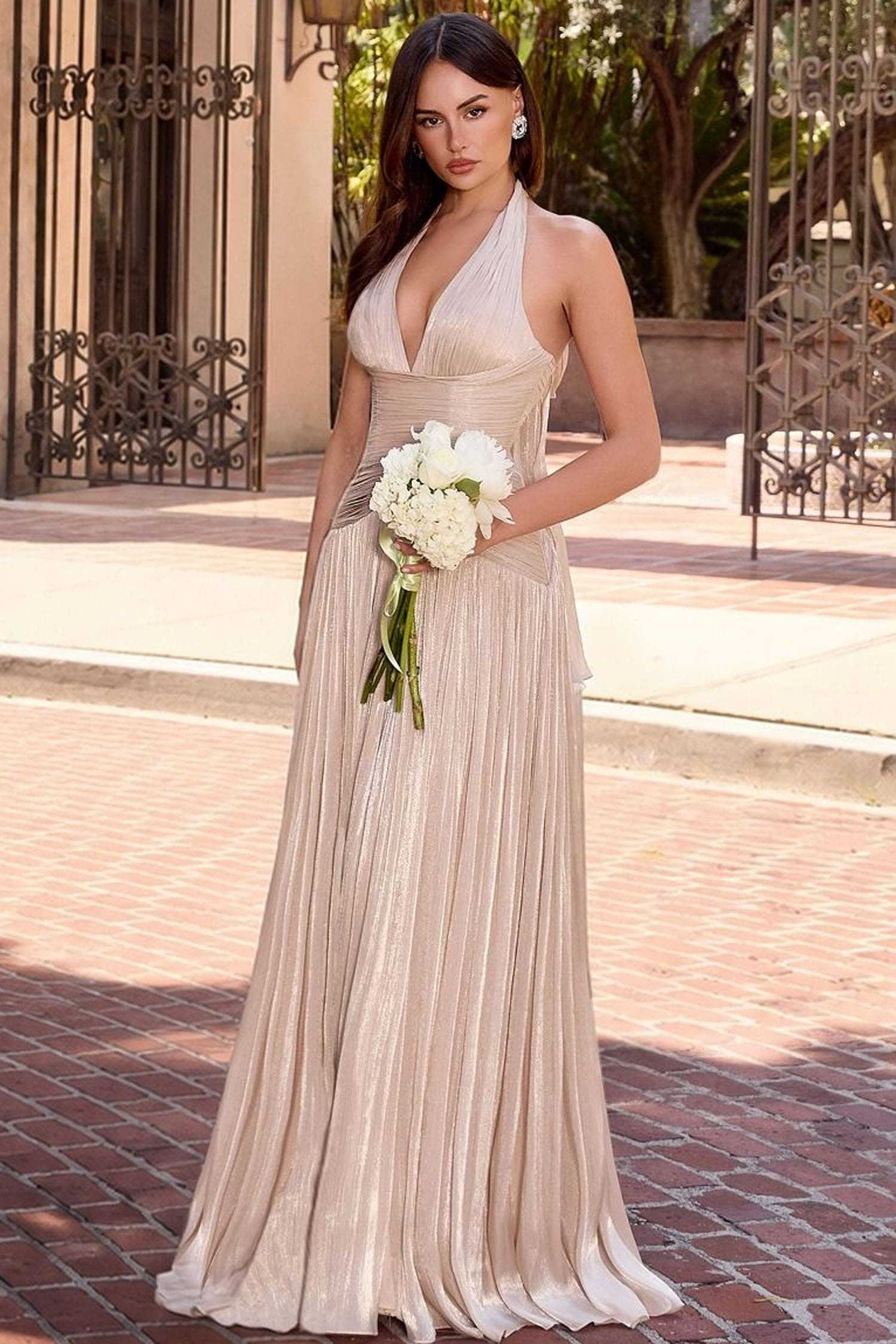 A woman stands outdoors on a brick walkway wearing a sleeveless, halter-neck, floor-length champagne pleated gown and holding a bouquet of white flowers.