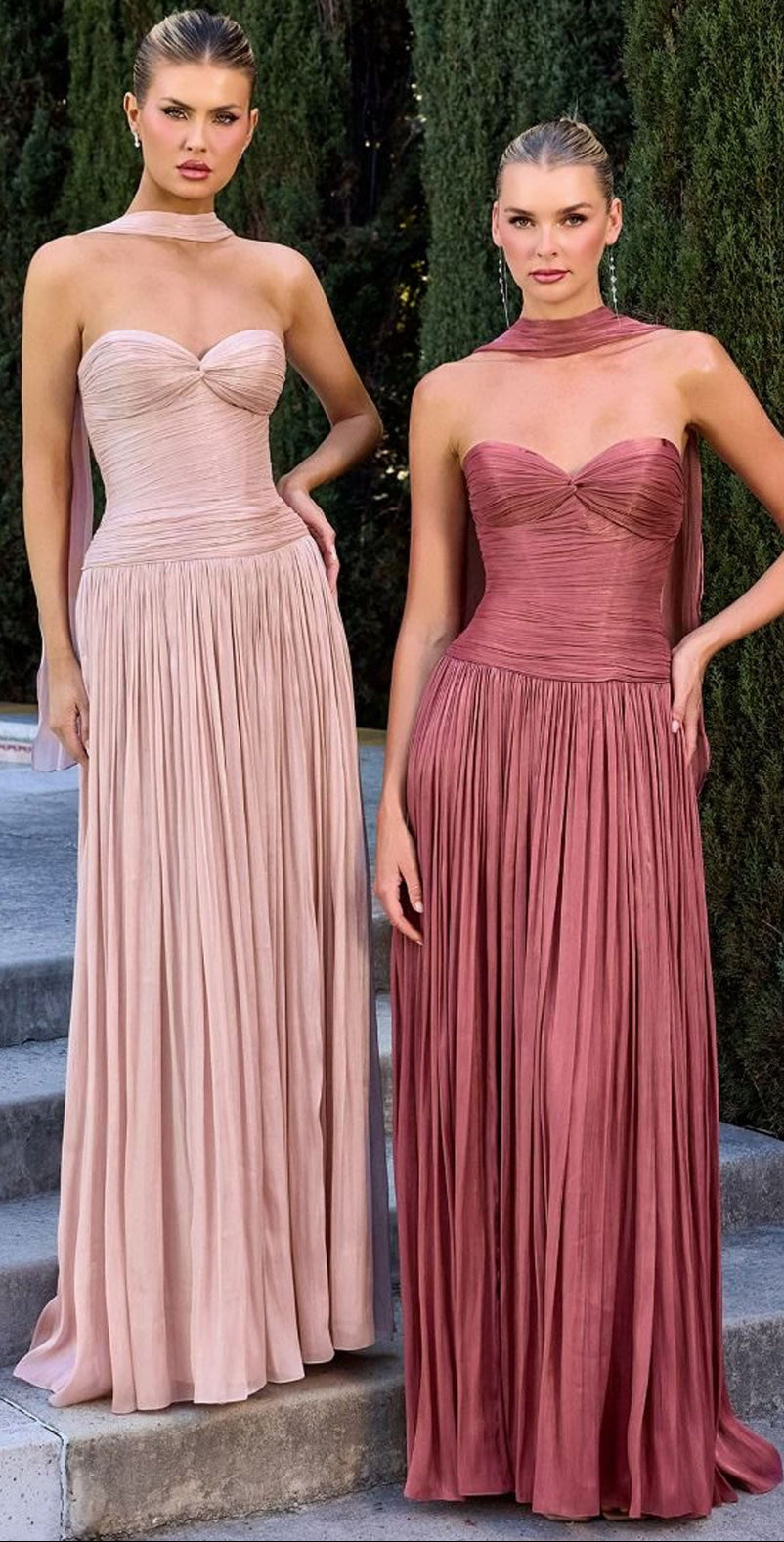 Two models wearing floor-length formal gowns standing on outdoor stone steps with tall green hedges behind them. The gown on the left is a light blush pink with a sweetheart neckline, structured bodice, and full pleated skirt, styled with a matching choker.