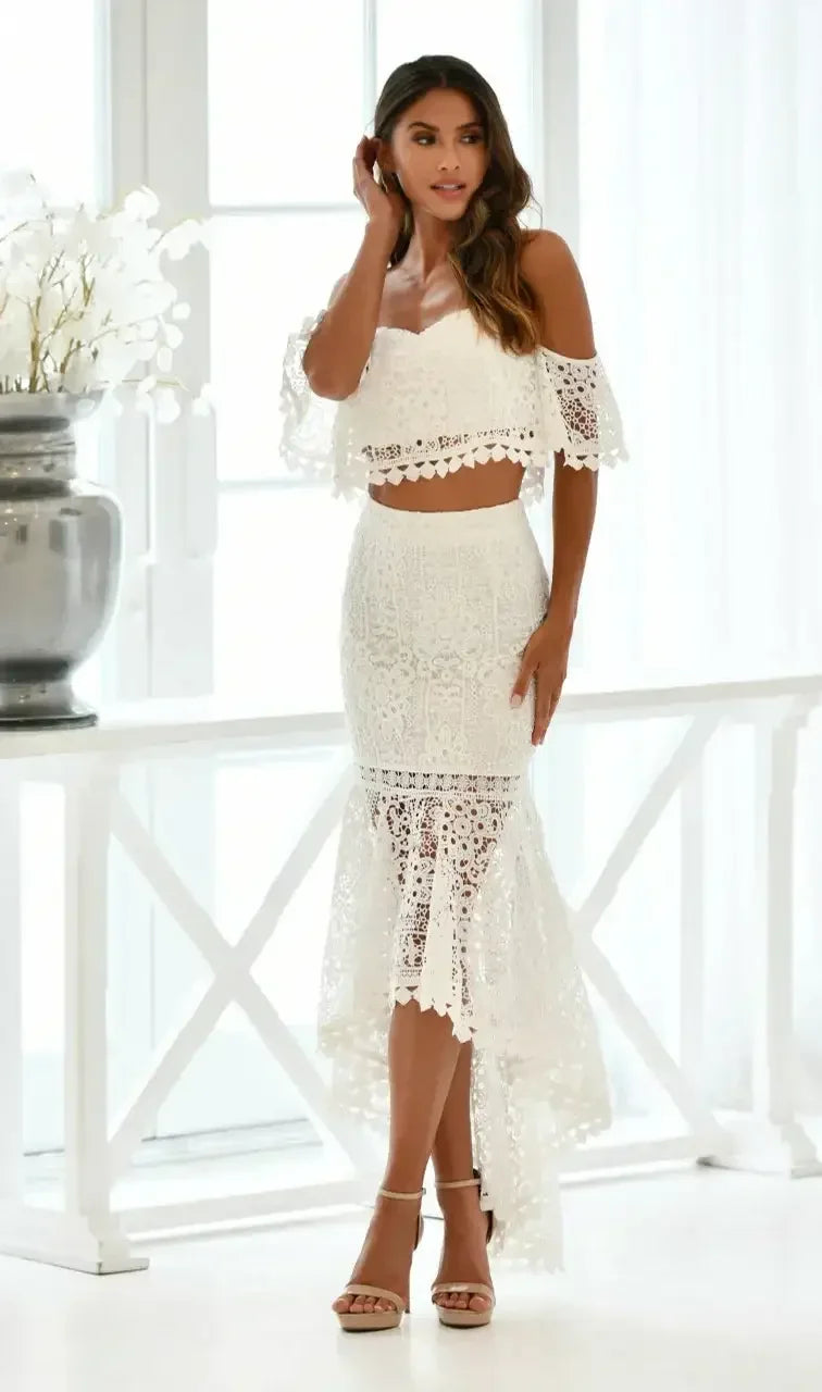 A woman stands gracefully in an elegant white lace two-piece outfit, featuring an off-shoulder top and a long skirt, against a bright, airy background with large windows and decorative flowers.