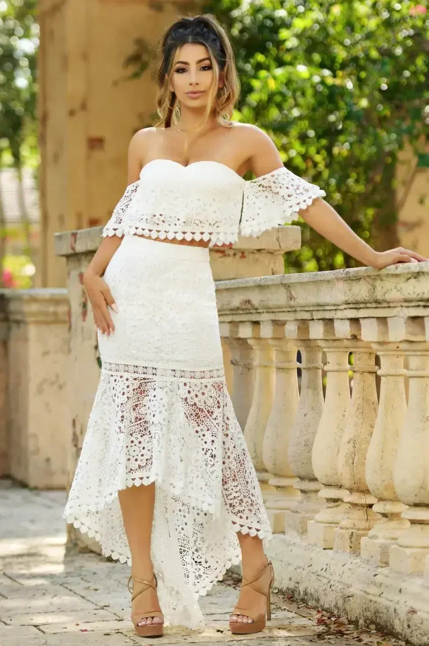 A woman stands elegantly by a stone railing, wearing a white lace two-piece outfit with a flared skirt. The surrounding area is lush with greenery and softly lit.