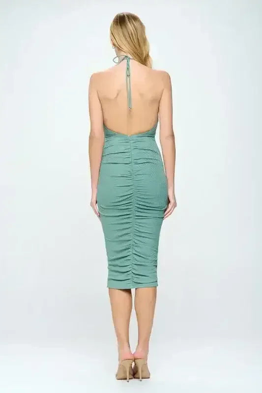 A fitted, green midi dress with ruching along the back is worn by a woman, showcasing her bare shoulders. The neutral background emphasizes the dress's color and design.