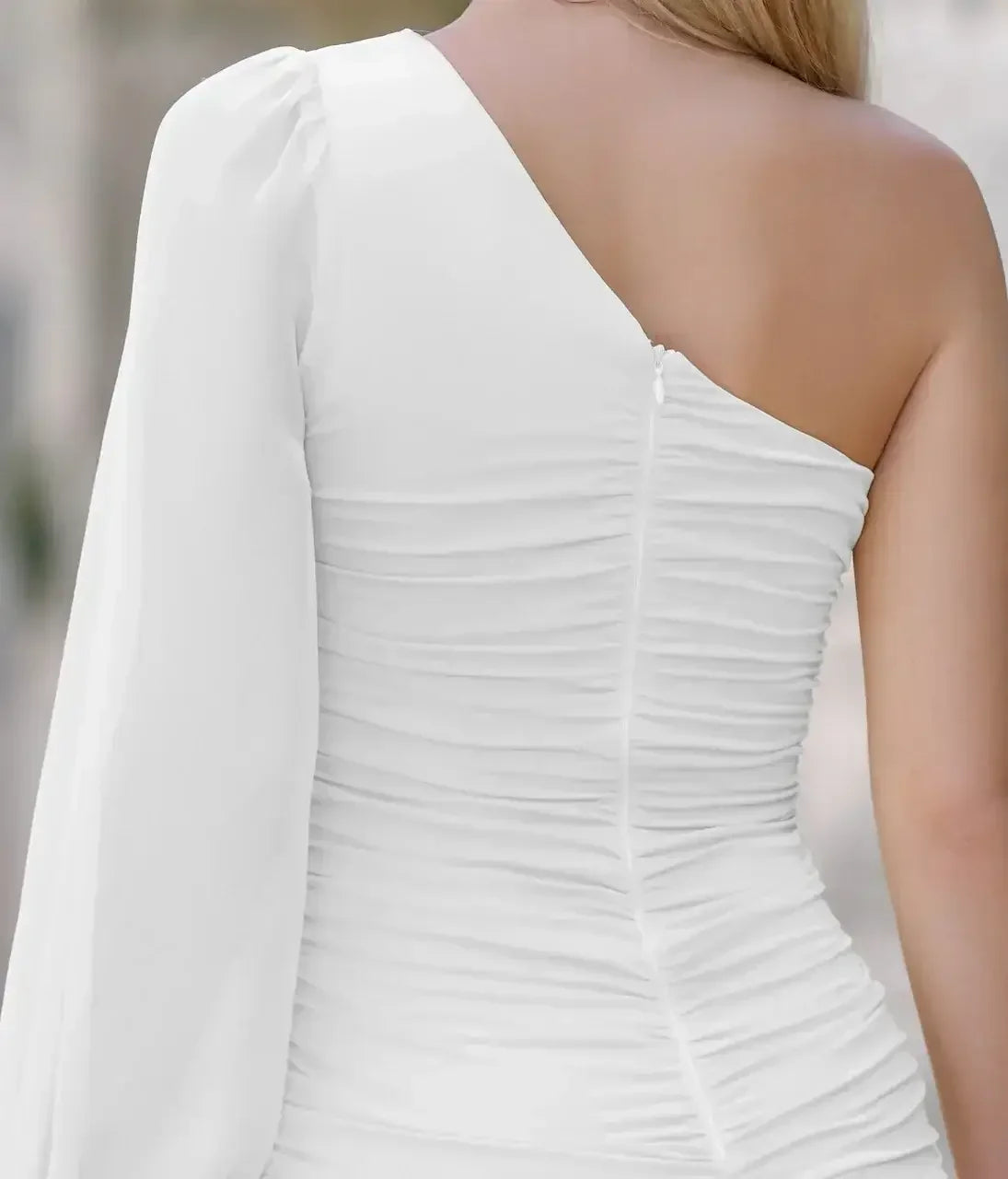 A fitted white dress with one sleeve features gathered fabric along the back and side, creating a textured appearance. The background is blurred, suggesting an outdoor setting.