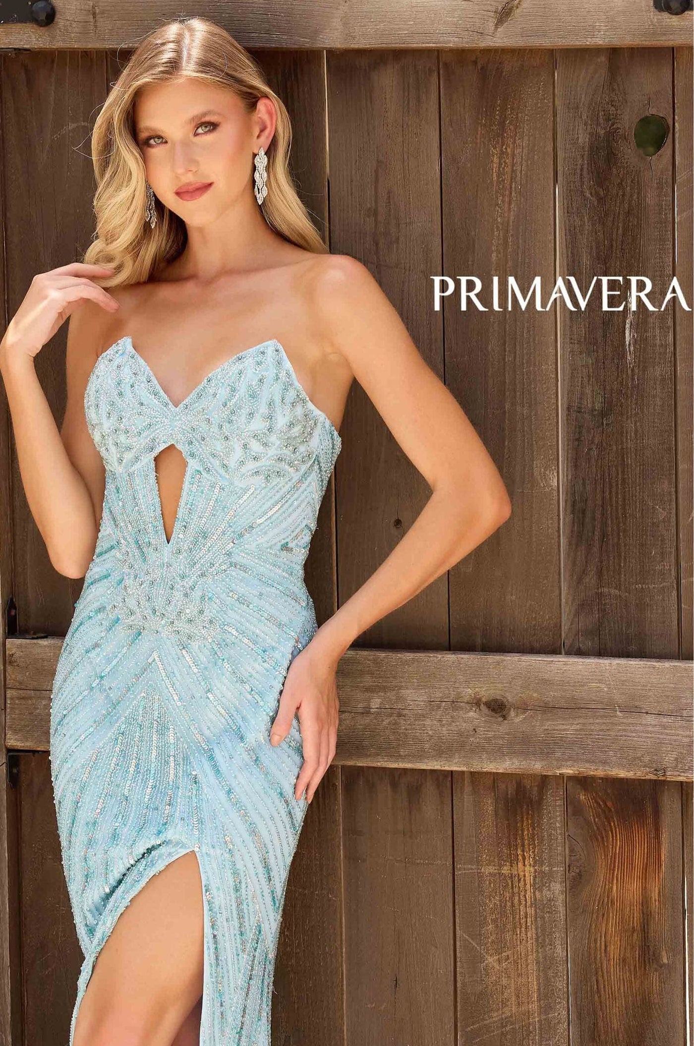 Woman wearing a light blue evening gown with a cutout design, standing against a wooden background with 'PRIMAVERA' branding.