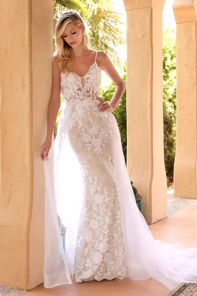 A woman stands gracefully in a delicate, floral lace wedding dress with a flowing overlay. She is posed against an architectural backdrop, surrounded by greenery and soft sunlight.