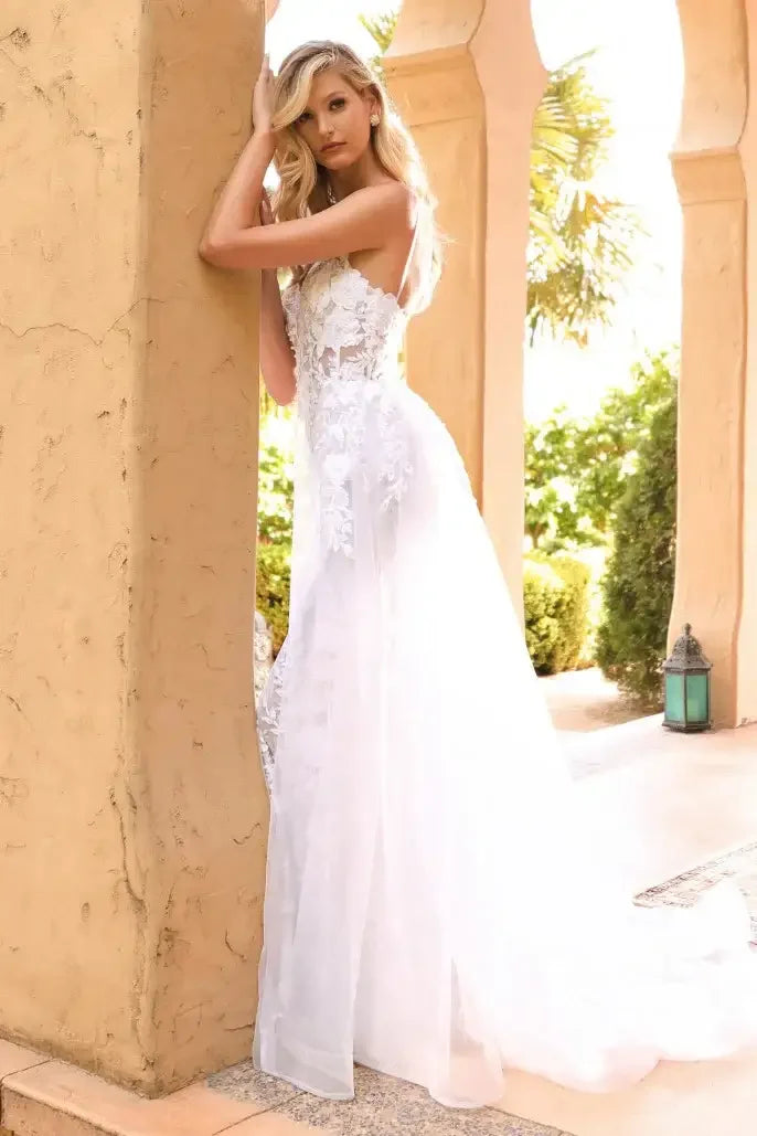 A woman in a delicate white gown leans against a textured beige pillar, with lush greenery and sunlight visible in the background, creating an elegant outdoor setting.