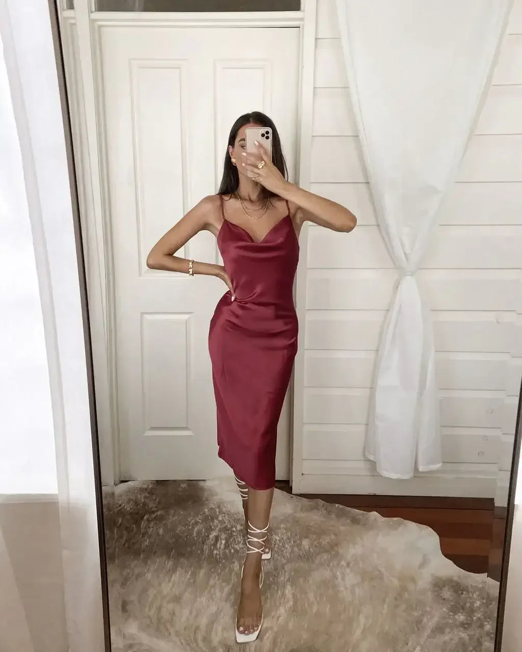 A woman poses in a fitted burgundy satin dress, holding a smartphone to take a selfie. She stands on a light-colored rug in a bright, minimalistic room with white curtains.